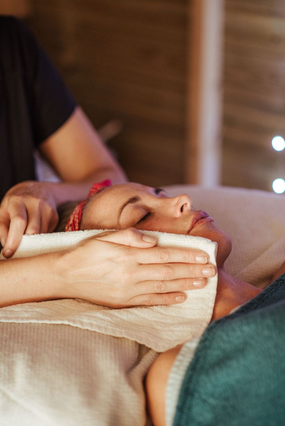A client resting on a treatment bed while an esthetician performs a facial treatment in warm light